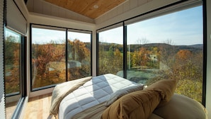 A bedroom with a large bed, a wooden ceiling, and large windows offering a view of autumn trees. - Amazing 34 foot high shipping container home in Whitingham on Sadawga Lake (Whitingham)