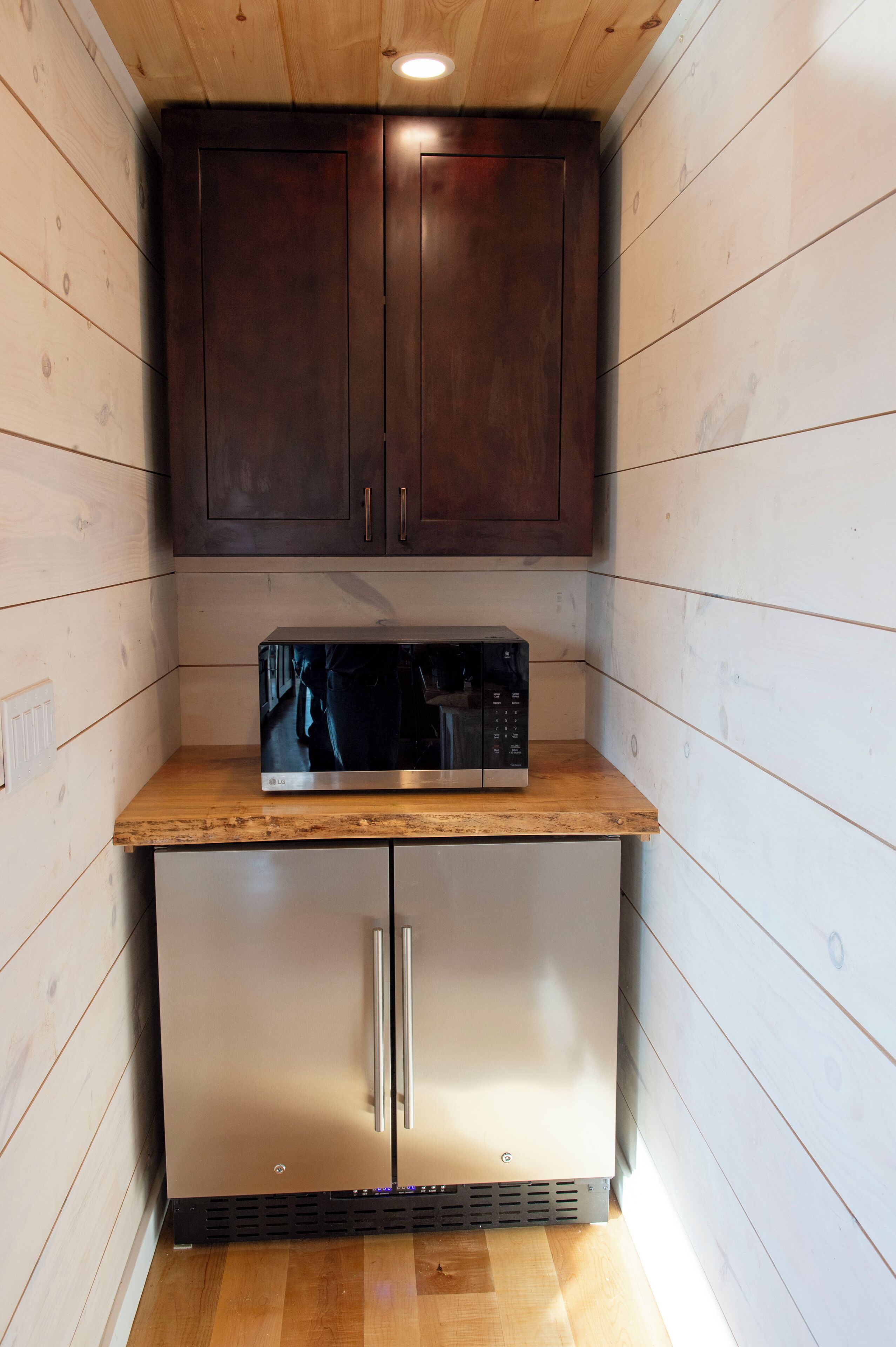 A compact kitchen with wooden walls, a microwave on a wooden countertop, and a stainless steel refrigerator.