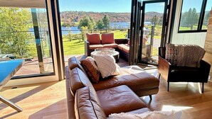 A modern living room with a large brown leather sofa, a small round table, and a chair. The room has large windows offering a view of a lake and trees. - Amazing 34 foot high shipping container home in Whitingham on Sadawga Lake (Whitingham)