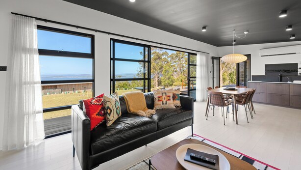 A modern living room with a black leather sofa, a dining table with chairs, and large windows offering a view of the outdoors. - The Outlook on Mount Louis - 2 Bedroom (Tinderbox)