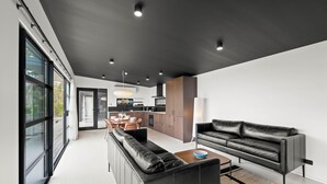 A modern living space with a black leather sofa, a dining area with a table and chairs, and a kitchen in the background. - The Outlook on Mount Louis - 1 Bedroom (Blackmans Bay)