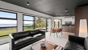A modern living room with a black leather sofa, a dining area with a wooden table and chairs, and large windows offering a view of the outdoors. - The Outlook on Mount Louis - 1 Bedroom (Blackmans Bay)