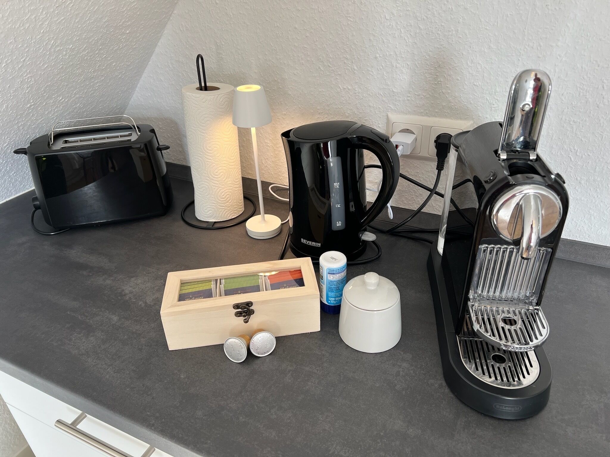 En köksbänk med en kaffebryggare, brödrost, pappershandduksställ och en trälåda med diverse föremål.
