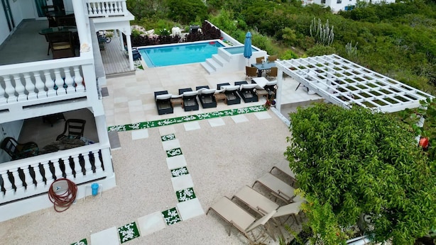 Un espace piscine aménagé sur le toit, avec des chaises longues et une pergola, entouré de verdure et offrant une vue sur un quartier résidentiel.