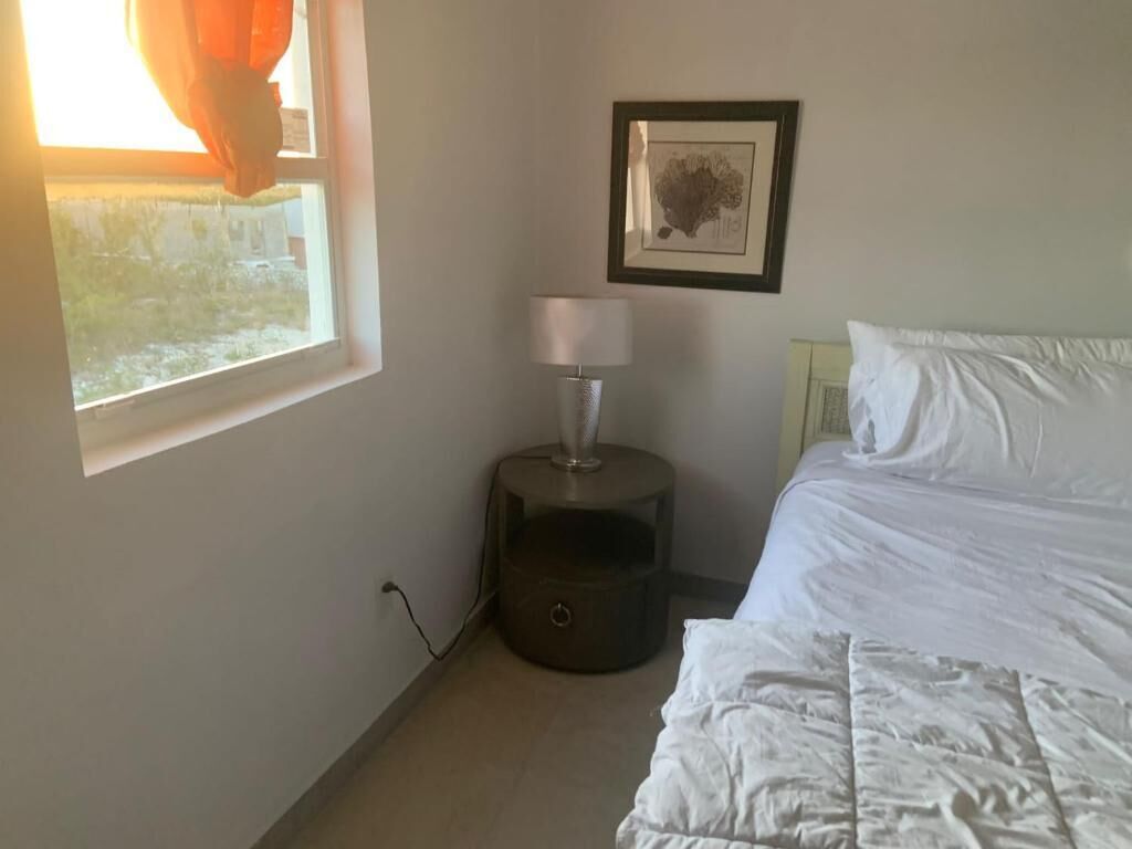 A bedroom with a bed, a bedside table with a lamp, a framed picture on the wall, and a window with curtains.