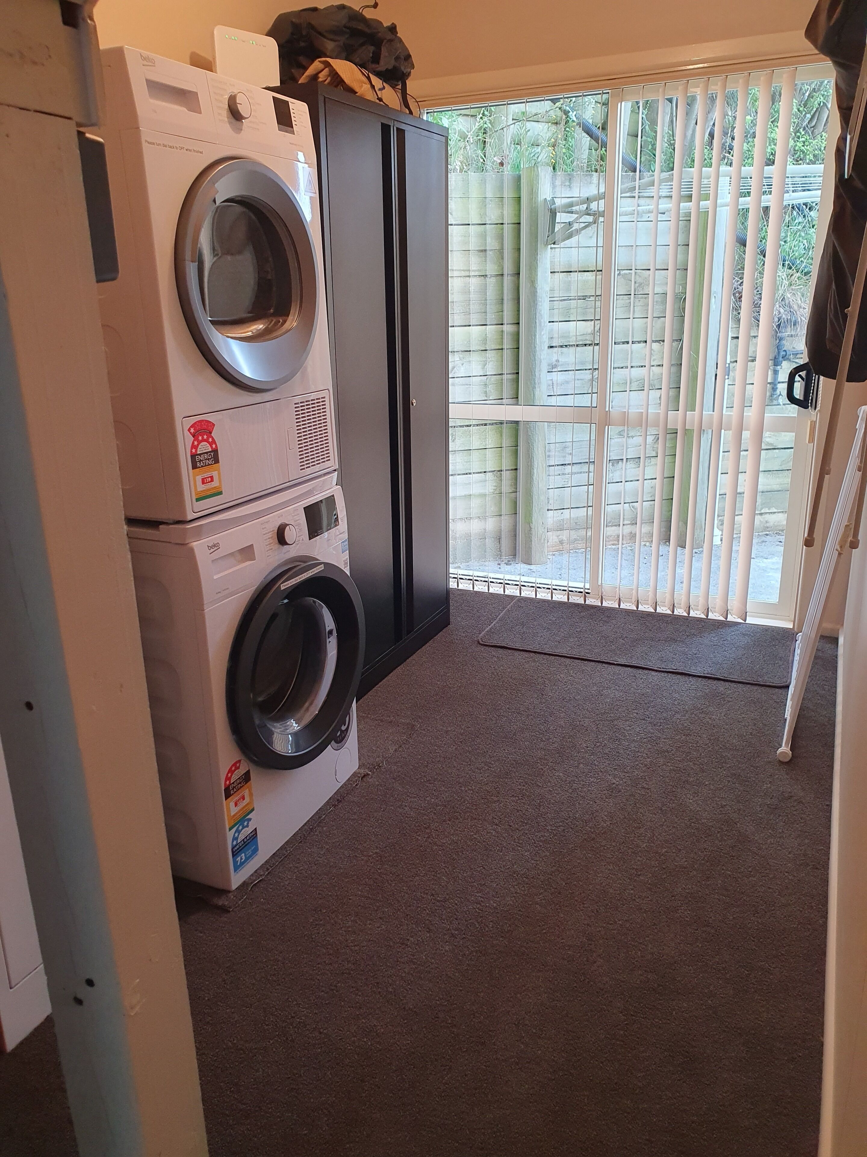 A laundry area with stacked washing machines and a dryer, a sliding glass door leading to a fenced outdoor area, and a carpeted floor.