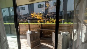 A view through a glass door of a wooden deck with wicker furniture, a wooden fence, and a building in the background. - Christchurch Central Apartment, Free Parking, 2-3 Beds (Christchurch)