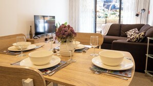 A dining area with a wooden table set for four, including bowls, plates, and wine glasses. A television is visible in the background, and a brown sofa with a patterned pillow is in the foreground. - Christchurch Central Apartment, Free Parking, 2-3 Beds (Christchurch)