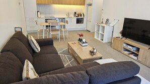 A modern living room with a sofa, coffee table, and TV stand. A kitchen with white cabinets and a dining area is visible in the background. - Christchurch Central Apartment, Free Parking, 2-3 Beds (Christchurch)