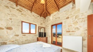 Un dormitorio con pared de piedra, techo de madera, cama con colcha estampada, cómoda, televisor y vista al mar a través de una puerta de cristal.