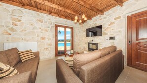Un salón acogedor con paredes de piedra, techo de madera y una chimenea. Hay dos sofás, un televisor y una puerta que da a un balcón con vistas al mar.