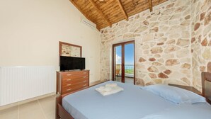 Un dormitorio con pared de piedra, techo de madera, cama con ropa de cama azul, televisor sobre un aparador de madera y un balcón con vistas.