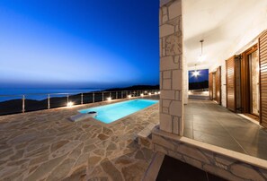 Una piscina de piedra con vista al mar al atardecer, un espacio al aire libre cubierto con puertas de madera y un muro de piedra.