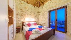 Une chambre avec un mur en pierre, un plafond en bois, un lit avec des oreillers et des serviettes, une table de chevet et une porte-fenêtre coulissante donnant sur un balcon.
