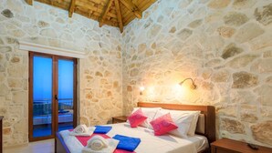 Une chambre avec des murs en pierre, un plafond en bois, un lit avec des oreillers blancs et roses, une table de chevet et un balcon avec une rambarde bleue.