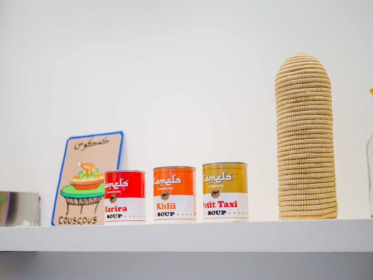 Three soup cans on a shelf with a sign and a tall, cylindrical object beside them.
