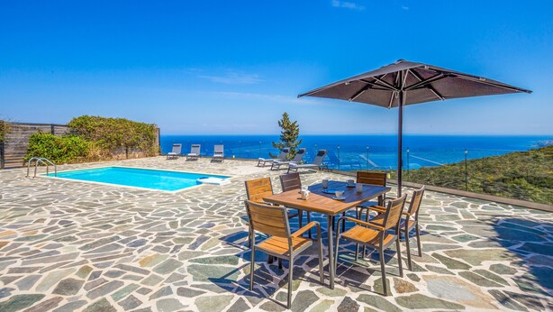 Una piscina in pietra con vista sull'oceano, un tavolo con sedie e un ombrellone.