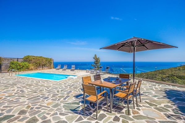 A stone pool with a view of the ocean, a table with chairs, and an umbrella.