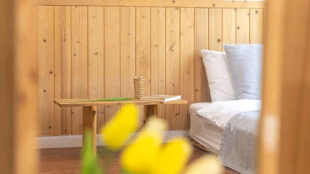 Een kamer met houten lambrisering, een bed, een kleine tafel met een boek en een glas, en een raam met gele bloemen.