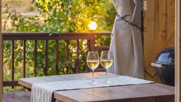 A wooden table with two glasses of white wine on a mat, outdoors with a wooden railing and greenery in the background.