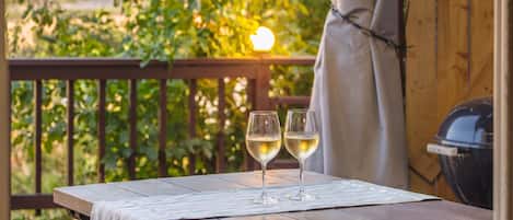 Una mesa de madera con dos copas de vino blanco sobre un tapete, al aire libre con una baranda de madera y follaje al fondo.
