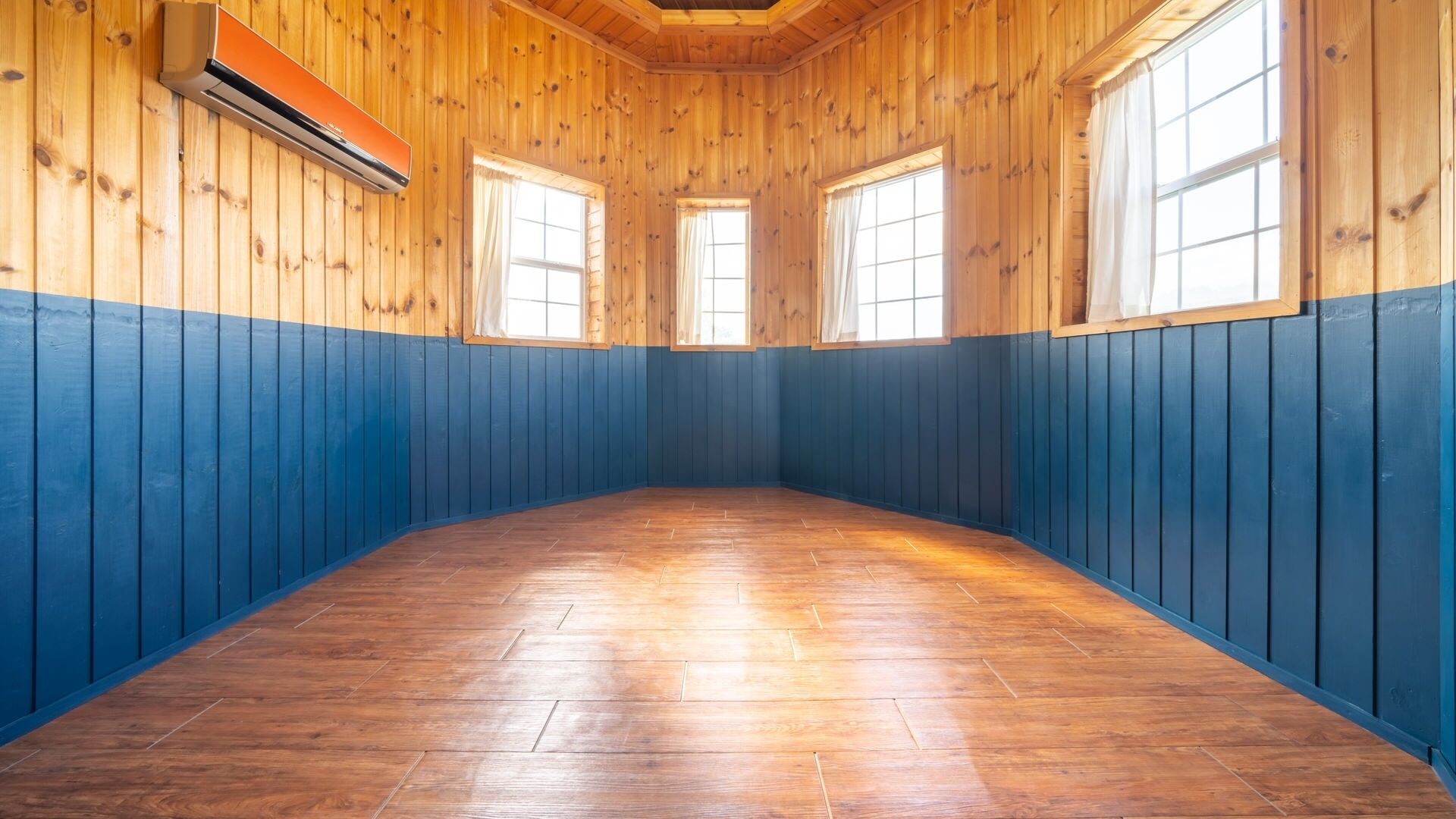 Quarto com paredes de madeira, parte inferior azul e piso de madeira. Possui três janelas com cortinas.