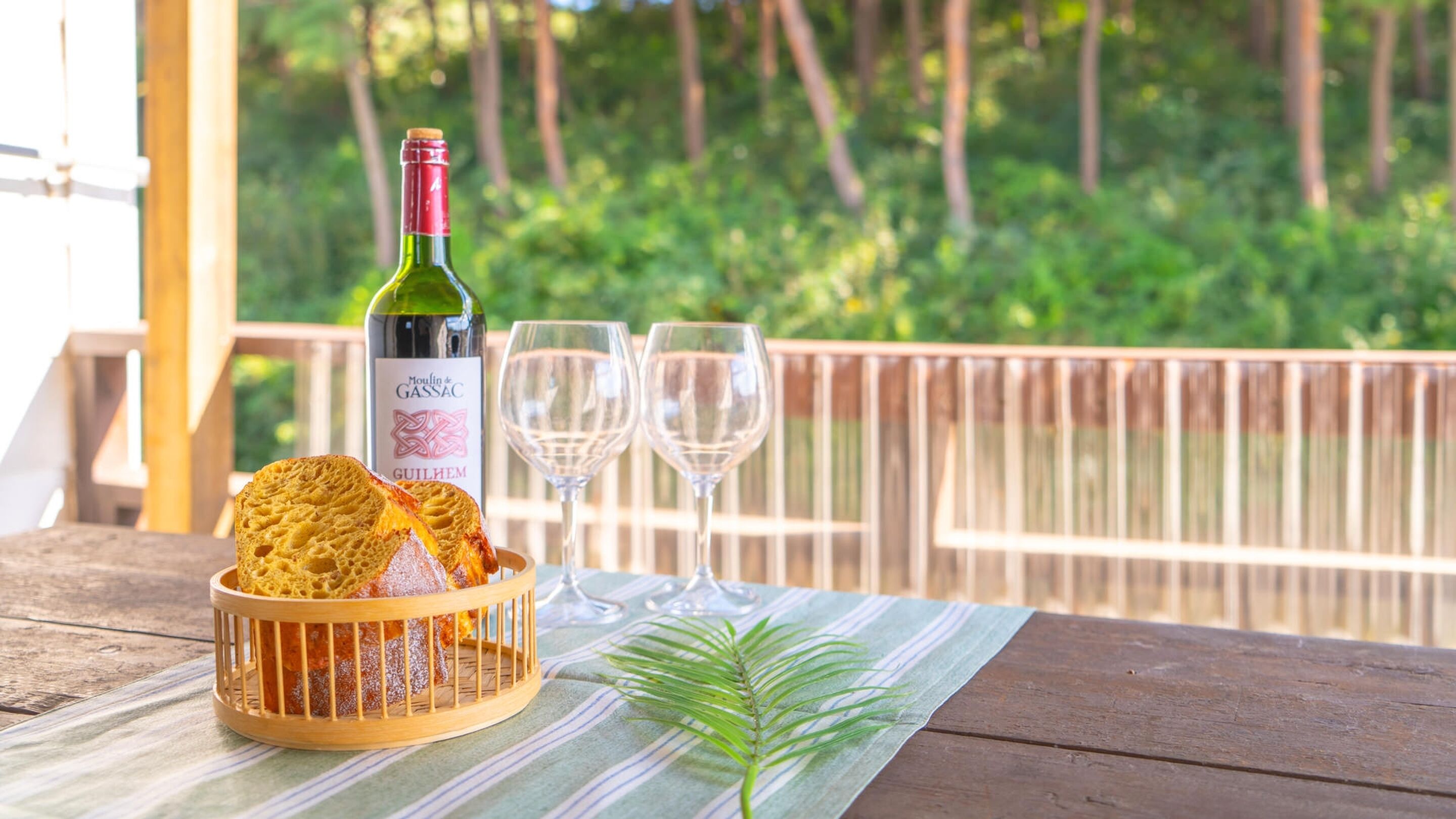 A wooden table with a bottle of wine, two glasses, and a basket of bread on a wooden deck with a view of trees.