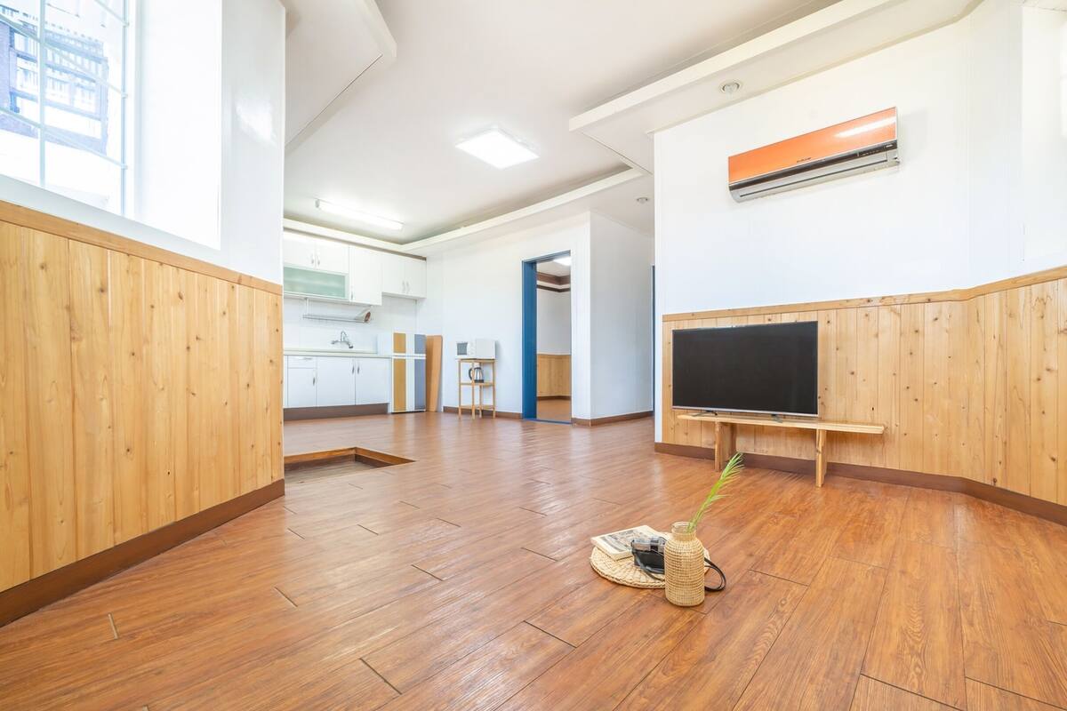 Una habitación amplia con suelo de madera, una kitchenette, una pizarra grande y un ventanal con vistas a edificios.