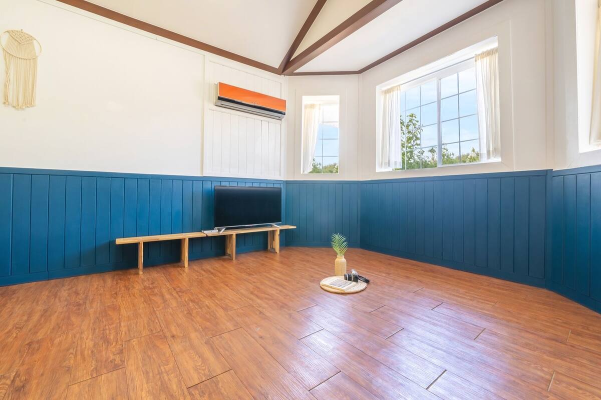 Una habitación con suelo de madera, un banco, un televisor y una ventana con cortinas.