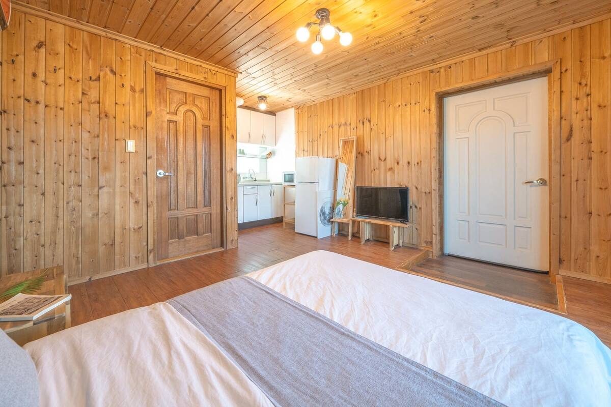 Habitación con paneles de madera, una cama, un televisor y una cocina visible a través de una puerta abierta.