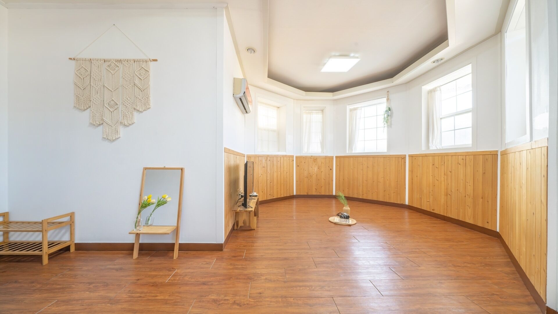 Een ruime kamer met houten lambrisering, een houten vloer, een macramé wandkleed, een houten plank met een spiegel en bloemen, en een klein tafeltje met een plant.
