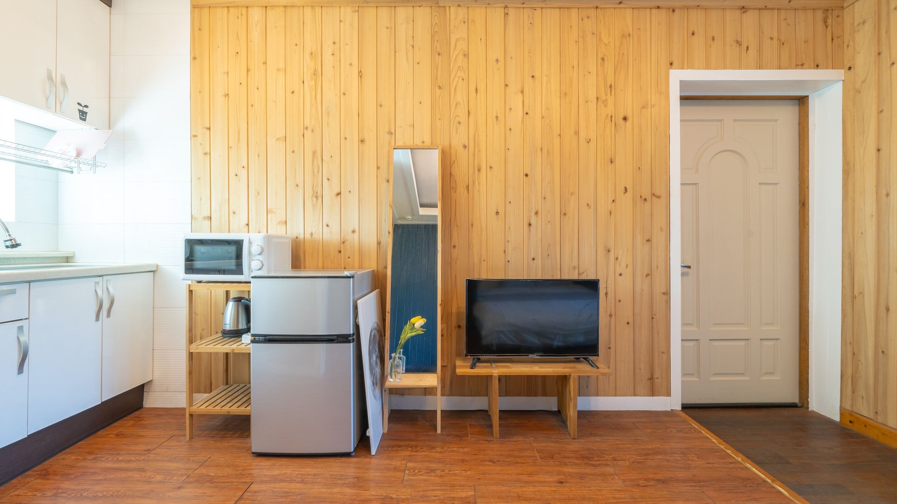 A compact kitchen with a refrigerator, microwave, and kettle, a small wooden table with a TV, and a door to the right.