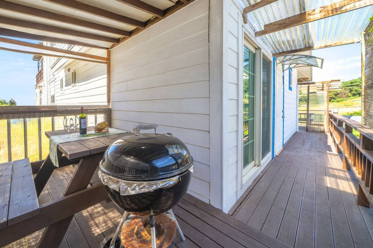 Un espacio al aire libre cubierto con una parrilla, una mesa con vasos y una botella, y vista a una casa.