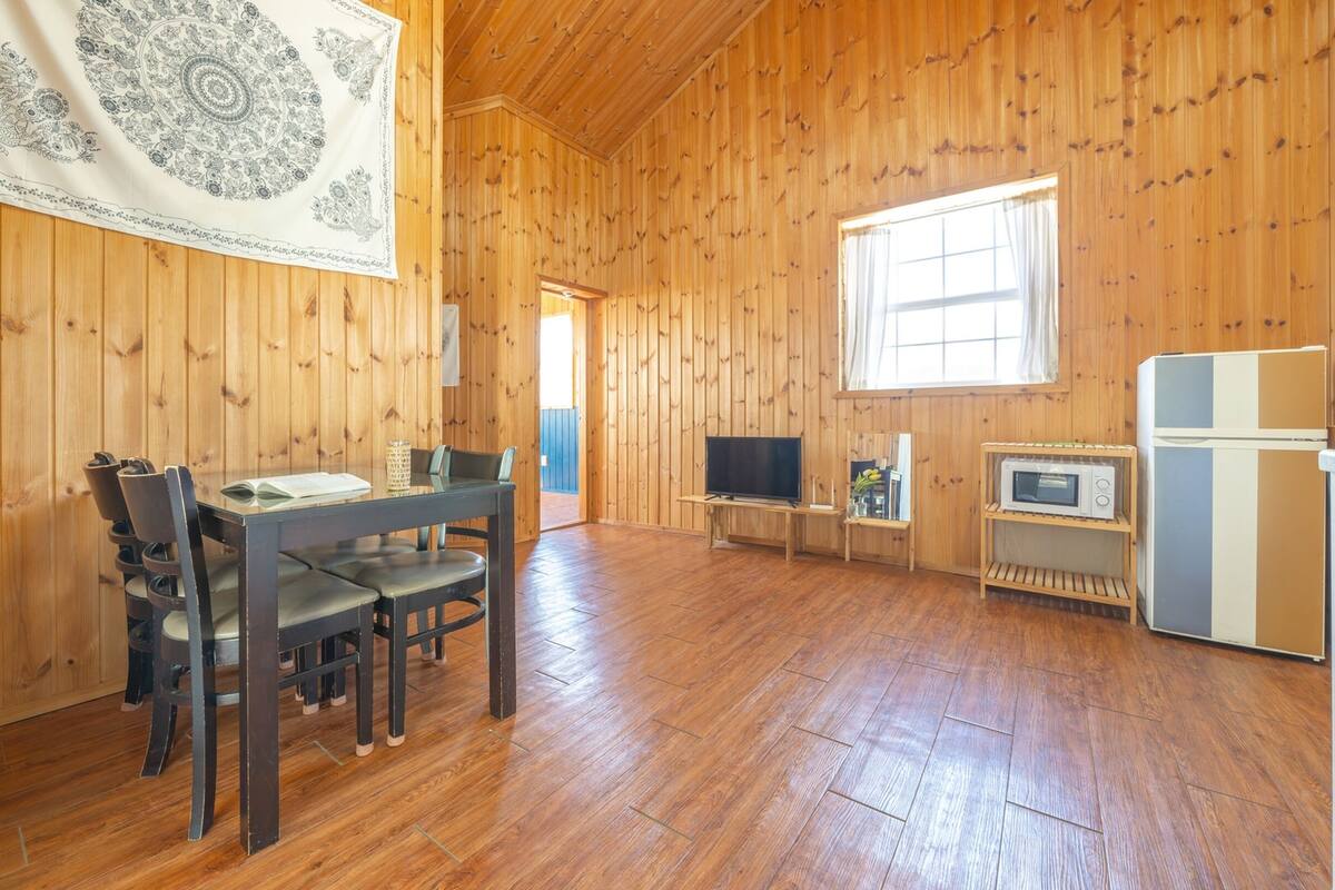 Una habitación con paneles de madera, una mesa de comedor, sillas, un televisor, un microondas y un refrigerador.