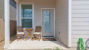 Eine überdachte Veranda mit zwei Holzstühlen und einem kleinen Tisch, eine Tür mit Fenster sowie eine grüne Pflanze auf der rechten Seite.