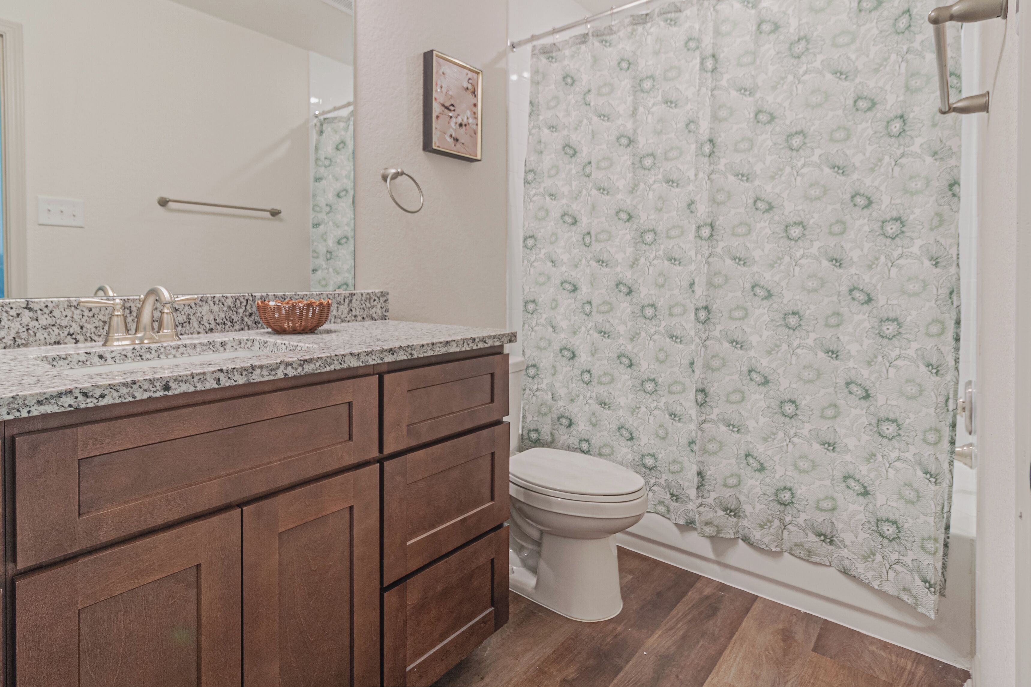 Un bagno con un piano in granito, armadietti in legno, un water bianco e una tenda da doccia con un motivo floreale.