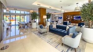 A spacious living area with a blue sofa, white armchairs, and a coffee table. There are potted plants, a large window with a view, and a television in the background. - Burj Khalifa Pool View Dubai Mall Address Opera 2B (Dubai)