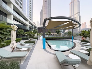 A rooftop swimming pool with lounge chairs and a covered seating area, surrounded by greenery and modern buildings.