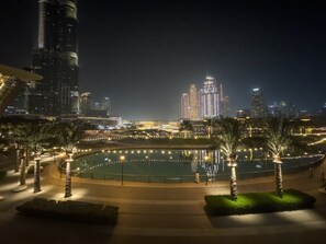 A nighttime view of a cityscape with illuminated skyscrapers, a circular water feature, and palm trees.