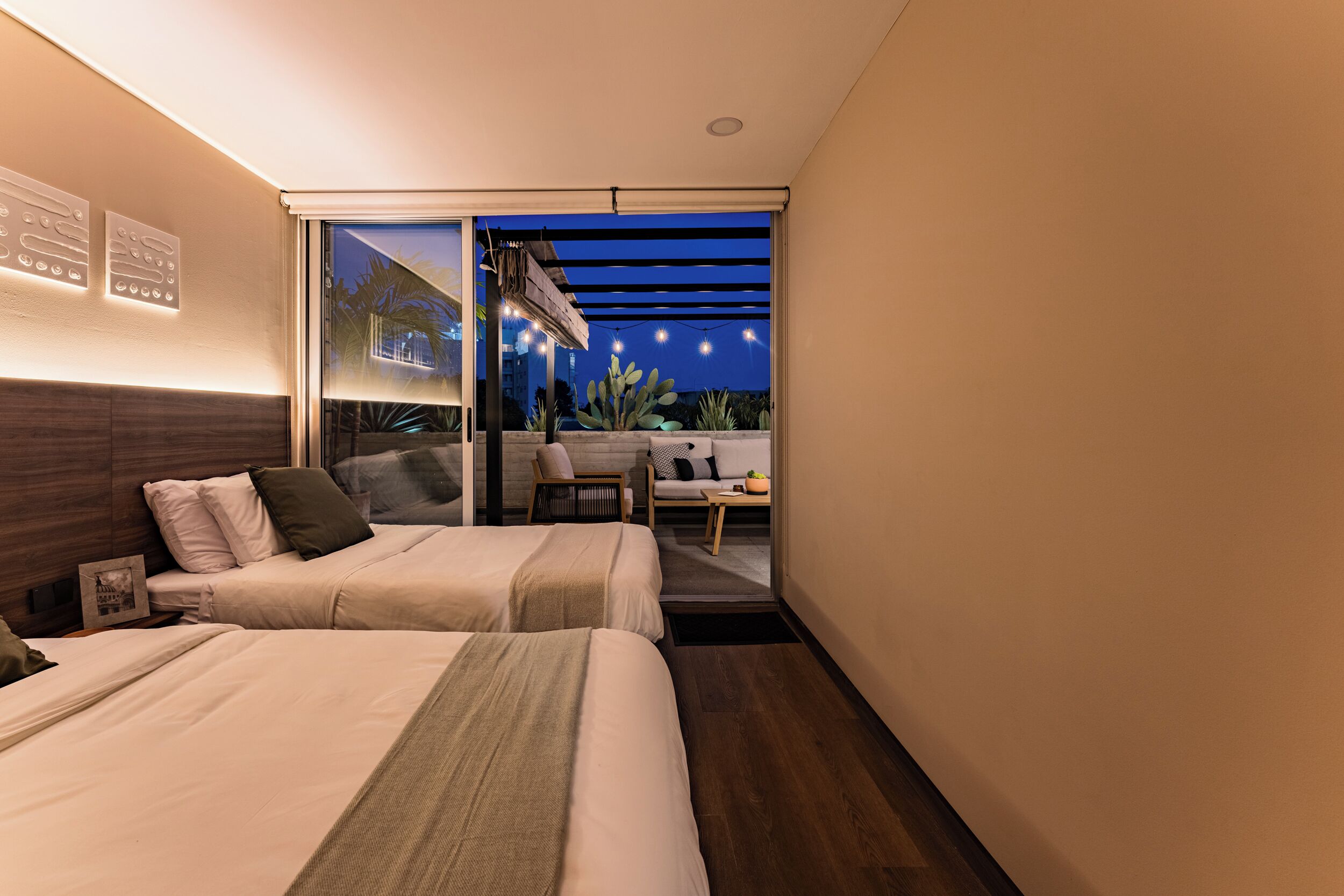 A modern hotel room with two beds, a balcony with string lights, and a view of the night sky.
