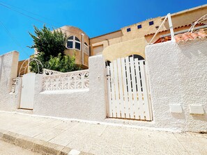 Uma propriedade murada com portão branco e paredes, entrada pavimentada e um edifício com telhado ladrilhado ao fundo.