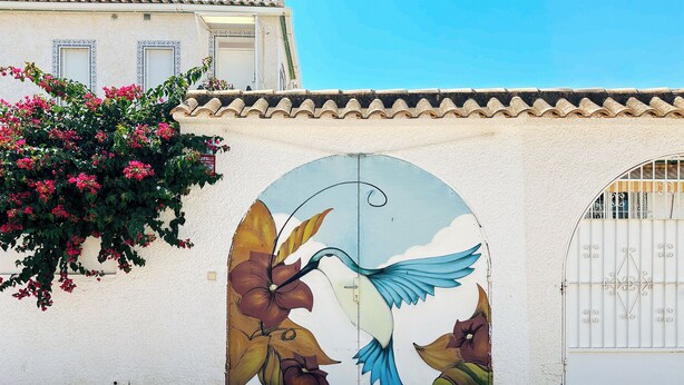 Un edificio con un mural de un ave y flores en la pared, una puerta blanca con reja de metal, y un tejado de tejas.