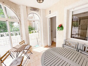 Quarto banhado pela luz do sol, com uma mesa redonda e dois bancos de madeira, uma porta branca e uma janela com vista para o verde.