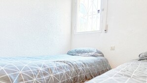 Un dormitorio con dos camas, una ventana con reja de metal y una pared blanca con textura.