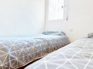 Quarto com duas camas, janela com grade de metal e parede branca com textura.