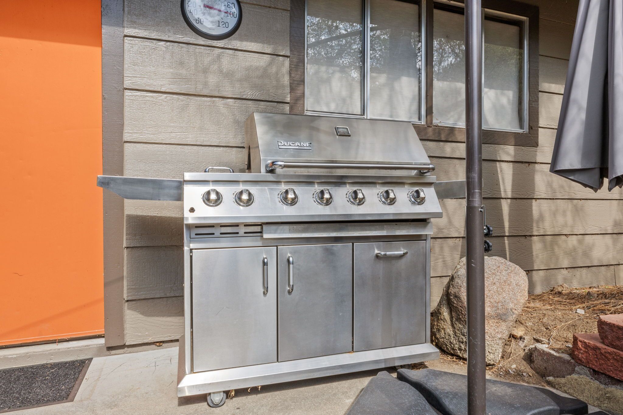 A stainless steel outdoor grill with four burners, a side burner, and a chimney, positioned against a wooden house with an orange wall and a window.