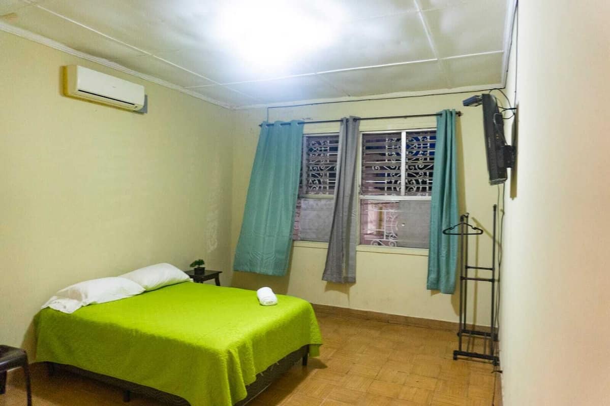 A small, single-bed room with a green bedspread, a window with curtains, a wall-mounted air conditioner, and a small table with a plant.