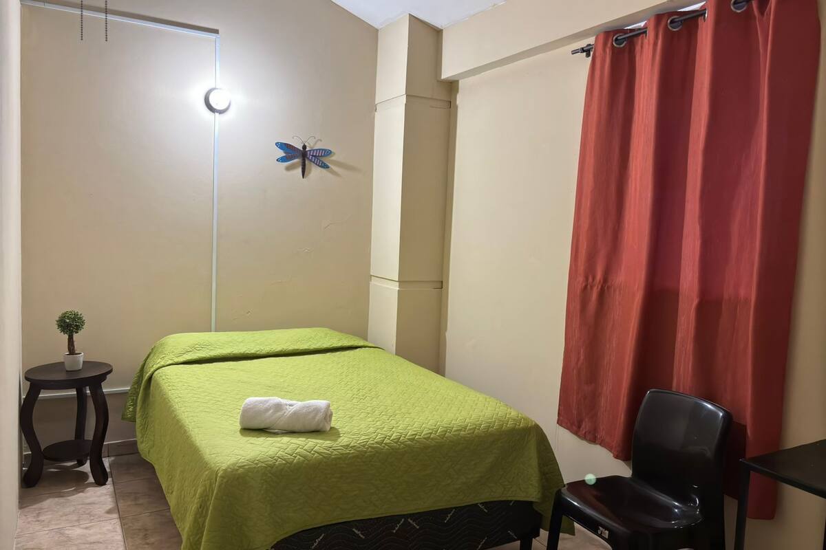 A small, neatly arranged bedroom with a bed, a chair, a small table, and a wall-mounted lamp.