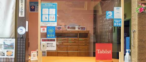 A hotel reception area with a 'Front' sign, a PayPay payment terminal, a 'Tabist' product display, and a wooden counter with drawers labeled with room numbers.
