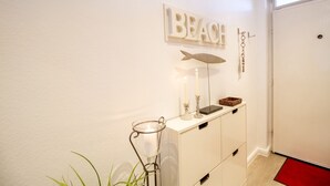 A hallway with a white wall, a 'BEACH' sign, a fish-shaped wall decoration, a small table with candles, and a red doormat.